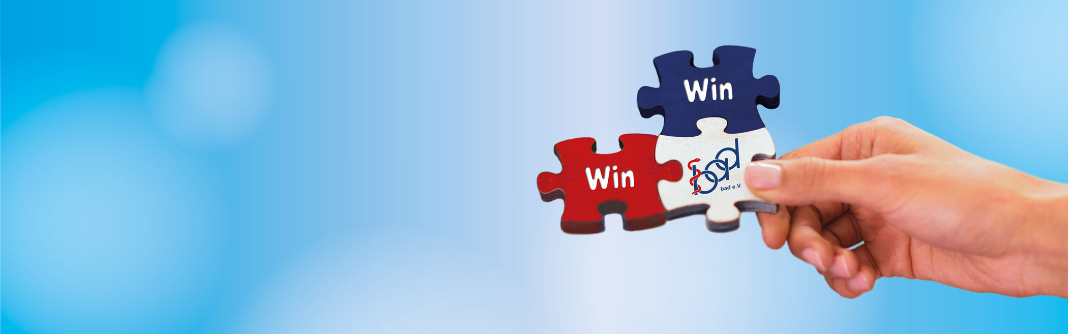 Bild eines Puzzles, das von einer Hand gehalten wird, mit einem blauen Himmel im Hintergrund. Die Puzzleteile sind rot, weiß und blau und tragen die Aufschriften 'Win' Win' sowie das Logo des bad e. V., das eine Verbindung zwischen den Elementen symbolisiert.