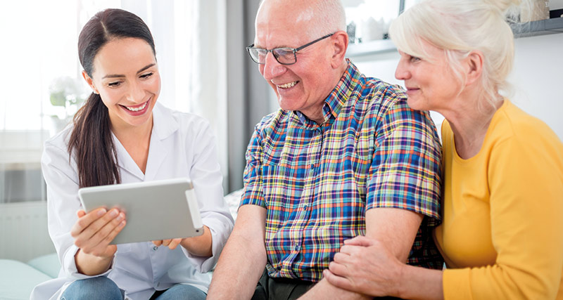 24 Qualifizierung zum Pflegeberater Ärztin schaut mit einem älteren Ehepaar lächeld auf Ihr Tablet.