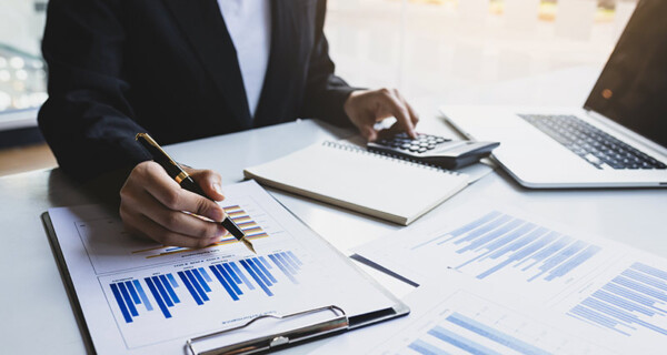 BWL-fuer-PDL_484441489_800x427 Eine Person tippt etwas in einen Taschenrechner und hält gleichzeitig einen Füller in der Hand, über einem Klemmbrett mit Grafen.